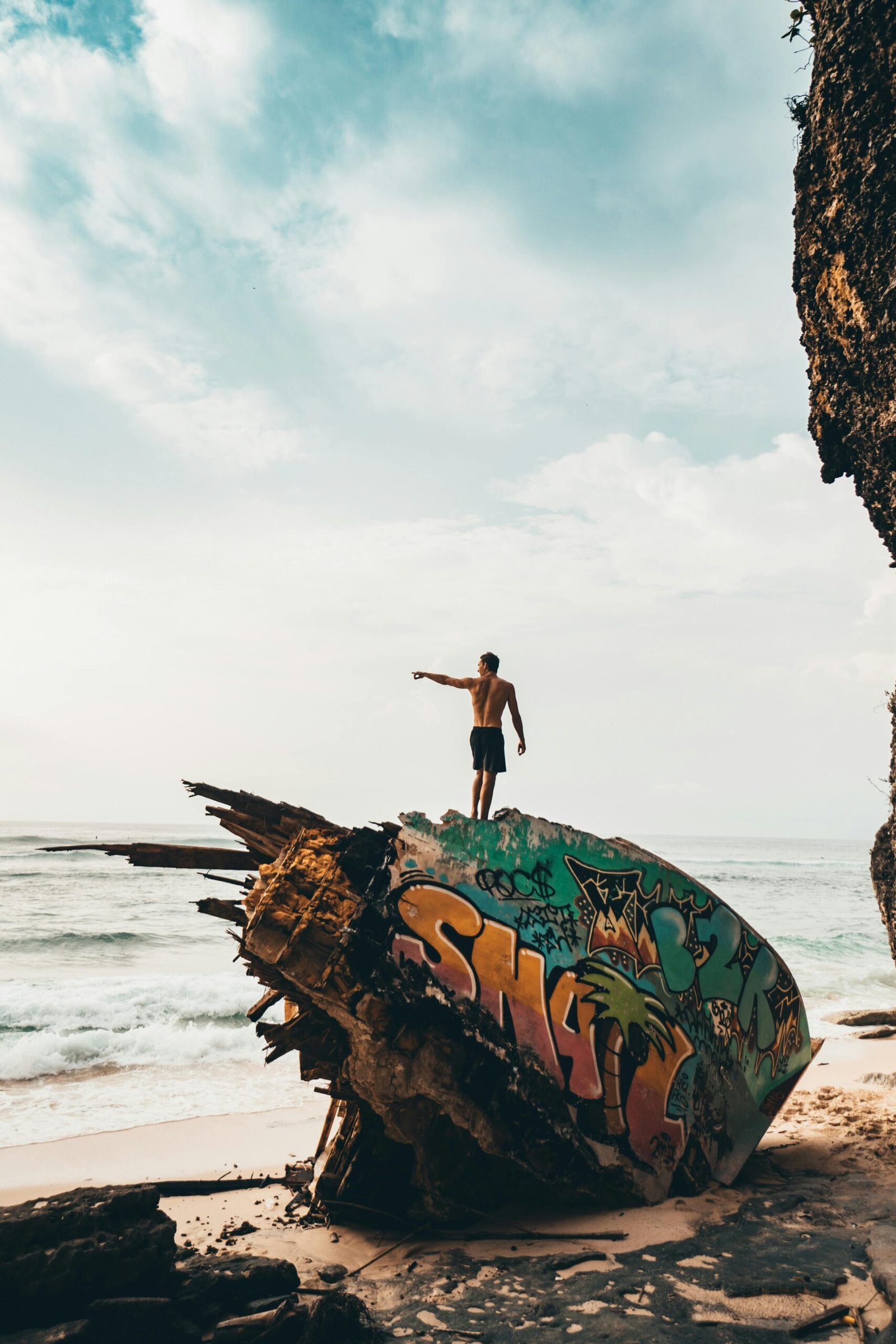 A man stands atop a graffiti-painted shipwreck on a beach in Bali, Indonesia, capturing moments of adventure and exploration.